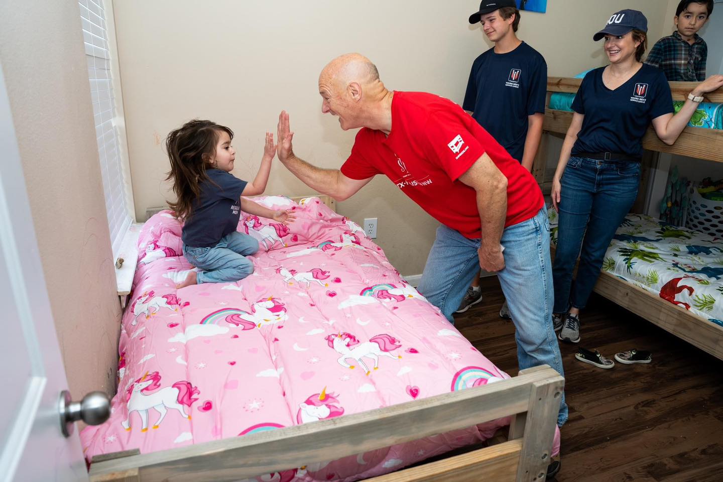 A group of three SHP volunteers delivers beds to children. A little girl sitting on her new bed with pink bedding gives a high-five to one of the volunteers.