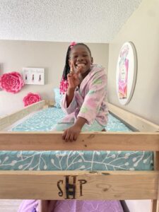 A group of three SHP volunteers delivers beds to children. A little girl sitting on her new bed with pink bedding gives a high-five to one of the volunteers.