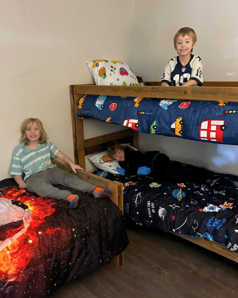 A boy sits on a twin bed with soccer themed bedding, while two other boys sit on the upper and lower parts of a bunkbed.