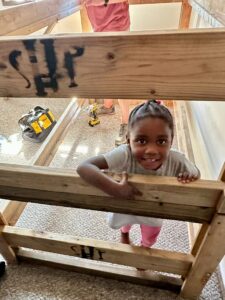 A little girl pokes her head through the wooden bunk bed frame that is being assembled in her home.