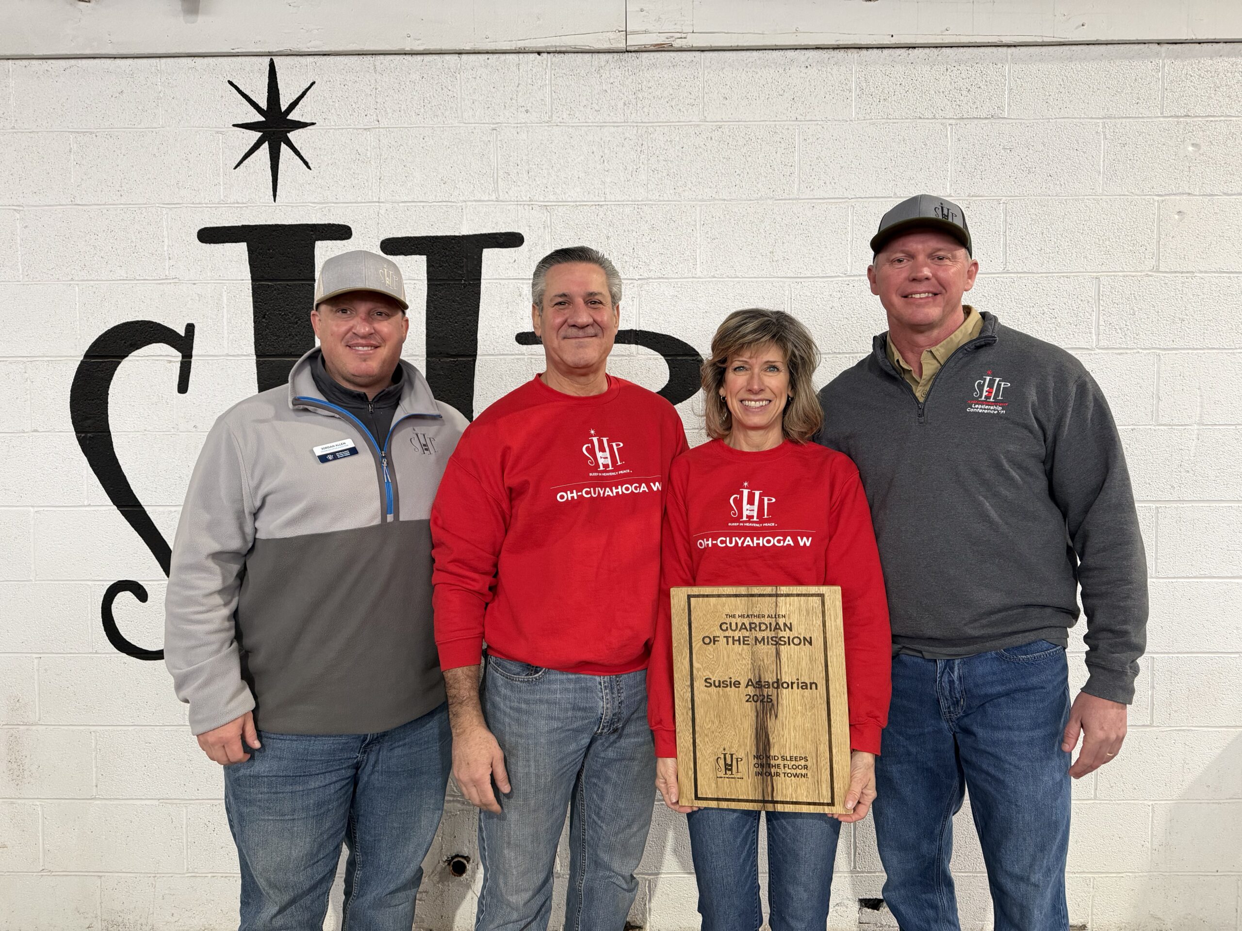 Image_20260221_093332 Four people stand together smiling in front of a white brick wall painted with the Sleep in Heavenly Peace logo. From left to right: Jordan Allen, SHP CEO, wearing a gray fleece; Kevan Asadorian in a red SHP OH-Cuyahoga W sweatshirt; Susie Asadorian, SHP-OH Cuyahoga W Chapter President, also in a red SHP OH-Cuyahoga W sweatshirt, holding a wooden plaque engraved with 'The Heather Allen Guardian of the Mission — Susie Asadorian, 2025'; and Eddie Arnold, Chair of the SHP Board of Directors, wearing a gray SHP pullover and cap.