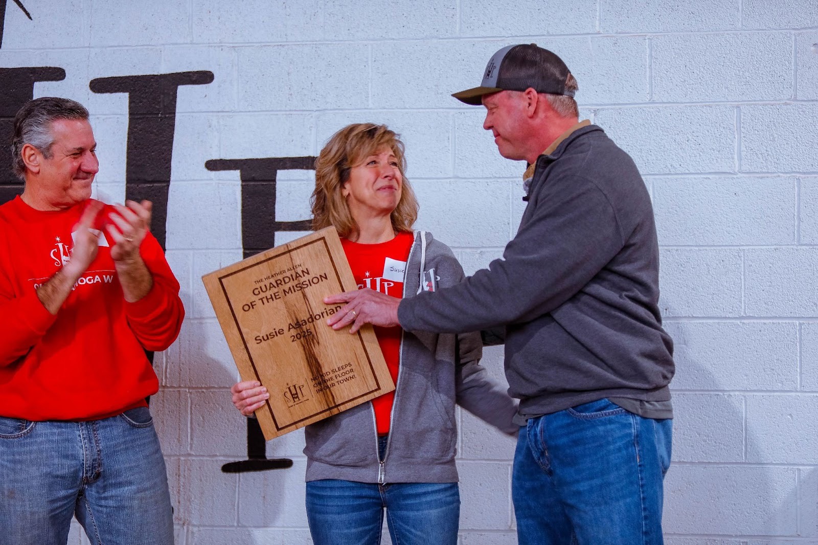 image Susie Asadorian stands in the center holding a wooden engraved plaque that reads "The Heather Allen Guardian of the Mission – Susie Asadorian 2025 – No Kid Sleeps on the Floor in Our Town!" Eddie Arnold, wearing a gray quarter-zip pullover and SHP cap presents the award to her while she reacts with visible emotion. Kevan Asadorian, in a red SHP sweatshirt, applauds to the left. The SHP logo is painted on the white cinder block wall behind them.