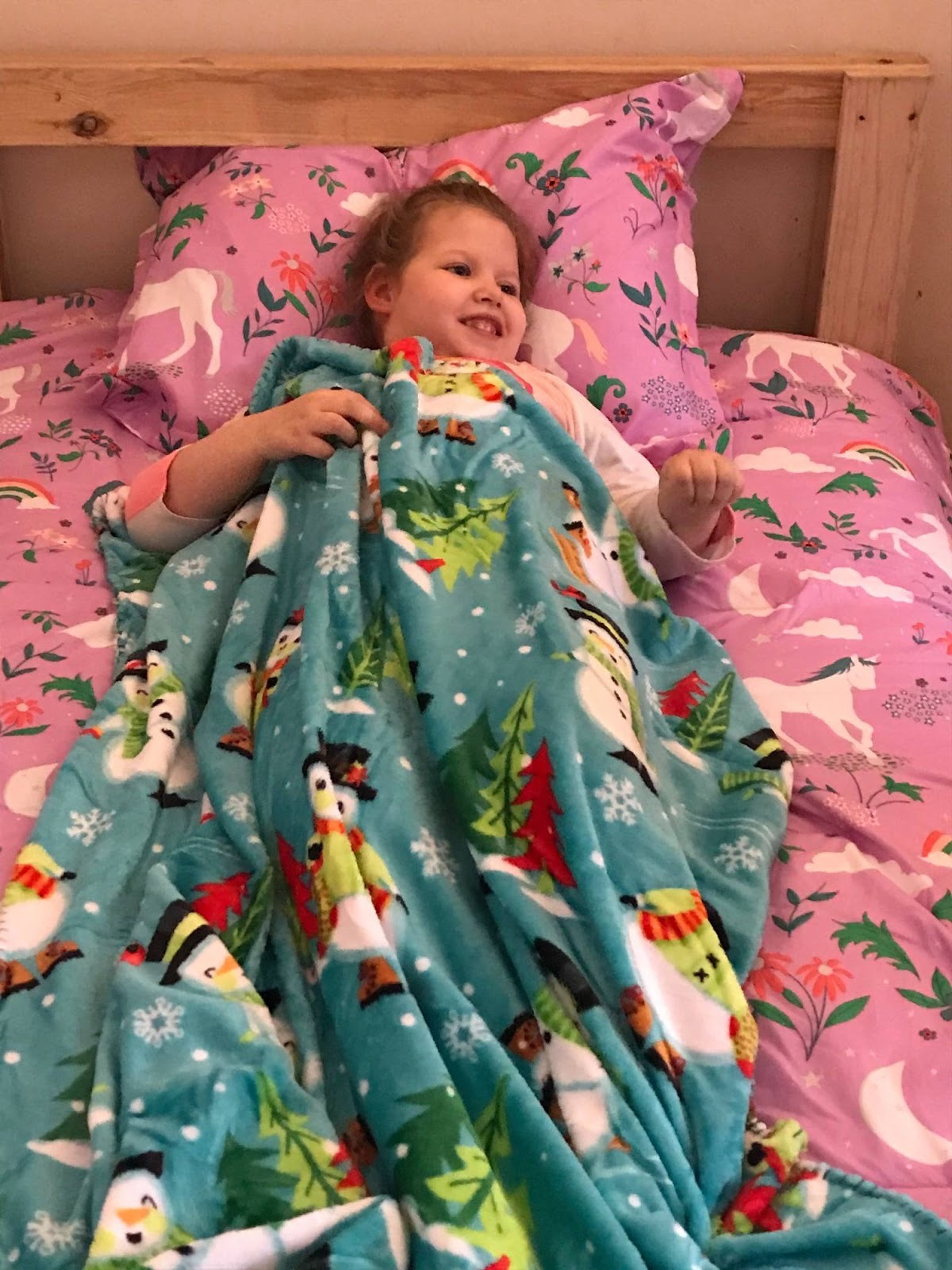 image A young girl beams with a big smile as she lies in her new SHP-built wooden bed, snuggled under a teal fleece blanket printed with snowmen and Christmas trees. She is surrounded by pink unicorn-print bedding and pillows. The natural wood headboard of her Sleep in Heavenly Peace bed frame is visible behind her.